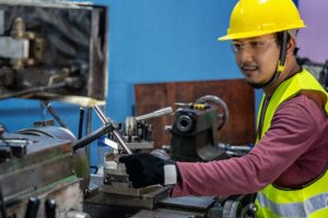 Asian machinist in safety suit operating the professional lathes in metalworking factory,