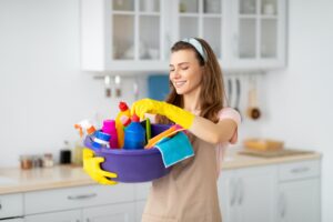 Portrait of positive young maid holding cleaning supplies at kitchen